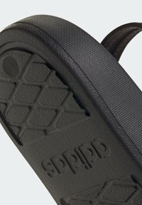 Černá podrážka žabky s texturovaným povrchem, obsahující geometrický vzorek běhounu a vyražené logo značky. Jednoduchý design pro pohodlí.