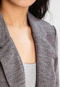 Szary, teksturowany blezer z kołnierzem szalowym, dopasowany przy szyi. Pod spodem widoczna gładka szara koszulka na ramiączkach. Subtelny wzór na tkaninie.