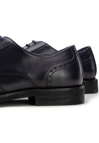 Czarne skórzane buty typu Oxford z ozdobnymi perforacjami na pięcie i grubymi, teksturowanymi podeszwami na białym tle.