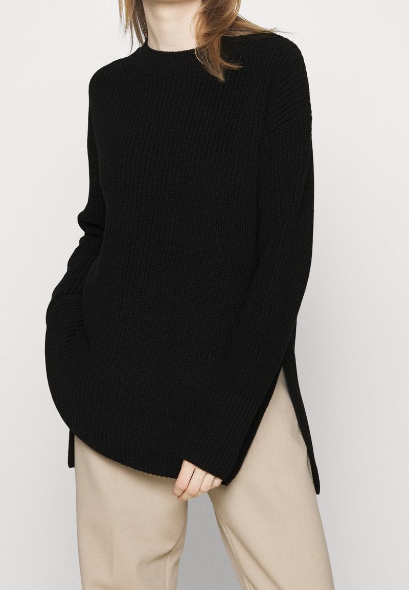 Femme portant un pull en maille côtelée noir avec des fentes latérales et un pantalon beige, debout devant un fond clair uni.