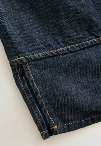 Der dunkelblaue Denimstoff weist eine sichtbare Textur mit hellblauen Einwebungen auf. Der Saum zeigt orangefarbene Nähte, die das Detail am Bündchen akzentuieren.