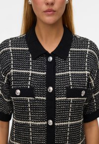 Ausu džemperis melnbaltā pārbaudes rakstā, ar melnu apkakli, īsām piedurknēm, sudraba pogām un divām priekšējām kabatām.