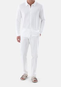 Biała, długorękawowa koszulka dzianinowa z teksturowanym wzorem, zapinana na guziki pod kołnierzykiem, zestawiona z białymi spodniami i jasnoszarymi klapkami na suwak.