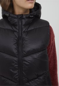 Svart pufferväst med huva, med dragkedjestängning och quiltad design. Bärs över en röd texturerad tröja.