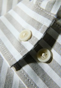 Camisa abotonada con rayas verticales en tonos marrón claro y blanco, con dos botones color crema y un acabado de tela texturizada.