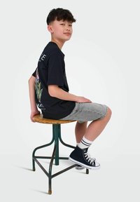 T-shirt noir à manches courtes avec un motif floral au dos, short en denim gris, et baskets montantes navy. Assis sur un tabouret en métal.