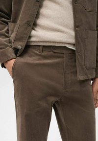 Chaqueta de sarga marrón con un acabado texturizado y pantalones a juego. Presenta botones oscuros y bolsillos laterales, llevada sobre un suéter claro.