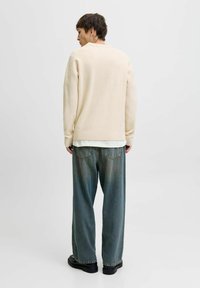 Cremefarvet striktrøje med ribbede manchetter, parret med løse, falmede blå jeans og sorte sko, set fra ryggen mod en ensfarvet baggrund.