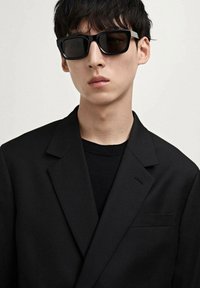 Black rectangular sunglasses with dark lenses, set on a model wearing a black suit jacket over a black shirt, featuring a textured finish.