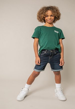 Un enfant aux cheveux bouclés se tient avec assurance, portant un t-shirt vert "Brésil", un short en jean retroussé, des chaussettes blanches et des baskets blanches sur un fond uni.