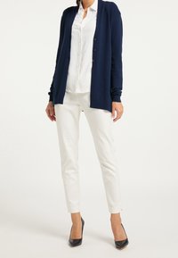 Marinblå cardigan med långa ärmar, öppen front, ribbat tyg. Kombinerad med en vit skjorta med knappar och vita slim fit-byxor, svarta högklackade skor.