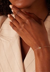 Gouden armband met een klein hartjesbedeltje aan een fijn schakeltje, gedragen aan een hand die nabij het gezicht is gepositioneerd, tegen een lichte stoffen achtergrond.