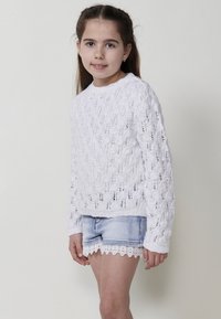 Pull en maille blanche avec un motif en gros grain, manches longues et col rond, associé à un short en denim bleu clair avec des bordures en dentelle.
