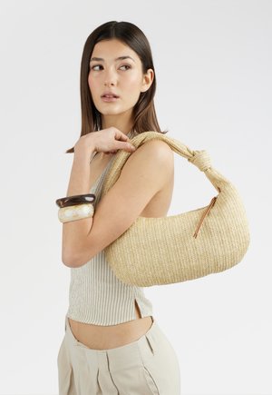 Donna con capelli lisci e scuri tiene una borsa beige intrecciata a spalla, indossa un top a righe senza maniche, pantaloni chiari e due braccialetti spessi.