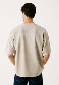 Beige übergroßes T-Shirt mit kurzen Ärmeln und Rundhalsausschnitt. Mit dezenten Druckgrafiken auf der Rückseite. Weicher, strukturierter Stoff.
