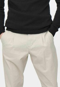 Un pull noir à côtes superposé sur un pantalon tailleur beige avec des plis sur le devant, mettant en valeur une texture lisse et une palette de couleurs neutres.