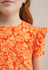 Apelsīnu krāsas t-krekls ar ziedu rakstu, ar volāniem uz piedurknēm, ribota tekstūra un apaļa kakla izgriezuma forma. Iezīmējas ar lielām, spilgtām ziedu ilustrācijām dažādās nokrāsās.