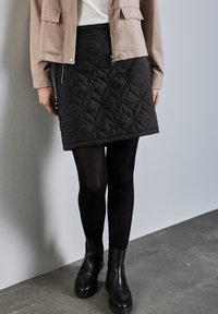 Schwarzer, gesteppten Minirock mit Reißverschluss und Seitentaschen, kombiniert mit schwarz blickdichten Strumpfhosen und Stiefeletten, vor einem grauen Hintergrund.