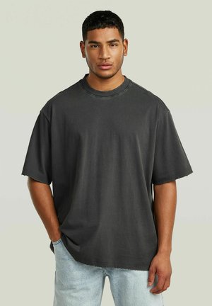 Jonge man met kort haar, gekleed in een loszittend, zwart vervaagd T-shirt en lichtblauwe spijkerbroek, staand met één hand in de zak tegen een effen achtergrond.