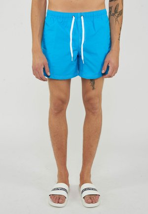 Uomo che indossa pantaloncini da bagno blu brillanti con coulisse bianche e sandali slide bianchi con testo nero, in piedi davanti a uno sfondo semplice.
