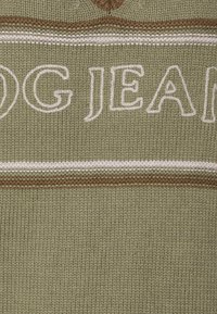 Olivově zelená pletená látka s bílými a hnědými horizontálními pruhy. Obsahuje text "DOG JEANS" v tučném bílém písmu. Texturovaný povrch.