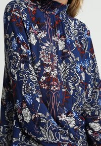 Långärmad blus i marinblått tyg med ett blommönster i vitt, rött och blått. Rynkad krage och lös passform med texturerad design.