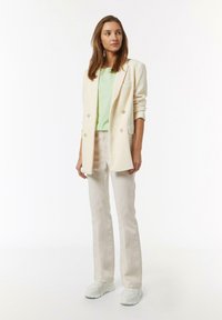 Light-colored, ribbed blazer with double buttons, paired with a green t-shirt and beige pants. White sneakers complete the outfit.