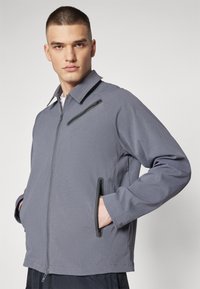 Gray zip-up jacket with a collared design, textured fabric, two zippered side pockets, and a subtle sheen on the surface.