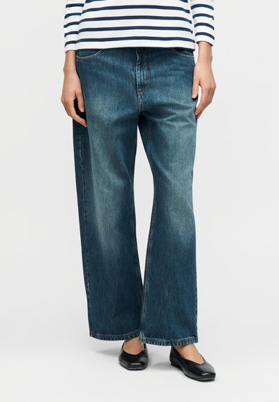 Jean en denim à jambes larges de couleur bleu moyen, avec un design classique à cinq poches et une texture subtilement délavée. Associé à des ballerines noires.