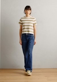 Maglietta cropped a righe beige e bianca abbinata a jeans blu. L'outfit è completato da sneaker chiare su un pavimento di legno.