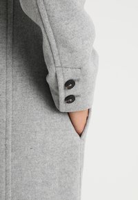 Mouw van een grijze wollen jas met een aansluitende pasvorm, twee donkere knopen en een zijzak, met een zachte textuur en strakke lijnen.