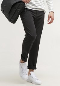 Black pants with folded cuffs, paired with white sneakers. The model holds a black shoulder bag and wears a striped long-sleeve top.