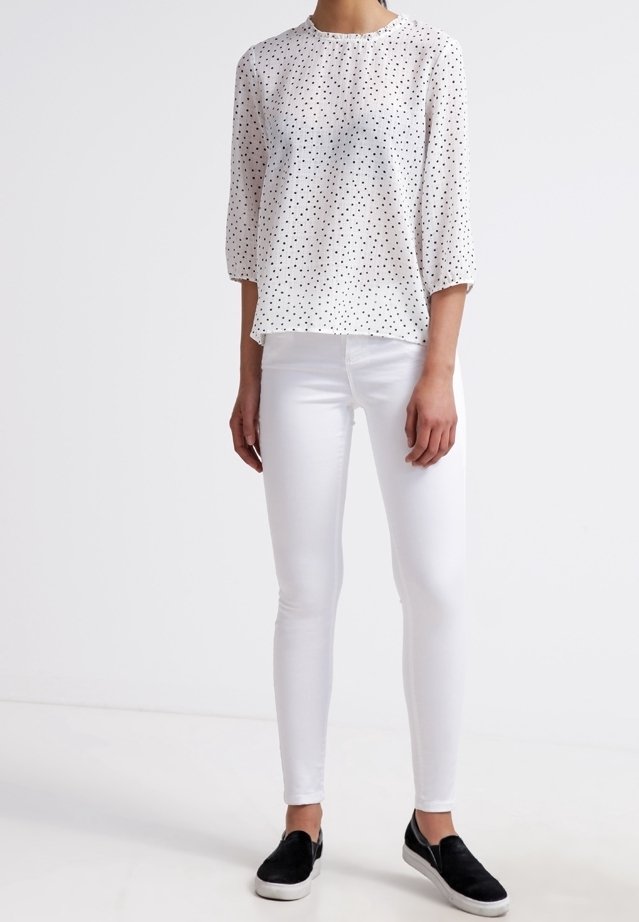 Blouse blanche à pois avec manches trois-quarts, associée à un jean skinny blanc et des chaussures à enfiler noires. Textures lisses et coupe décontractée.