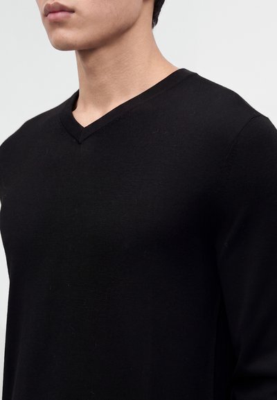 Jeune personne portant un t-shirt noir à manches longues avec un col en V, sur un fond uni gris clair visible.