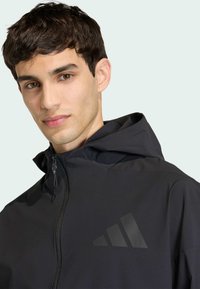 Giacca leggera nera con cappuccio, dotata di una cerniera frontale e un logo nero opaco sul petto. Texture liscia e design aderente.