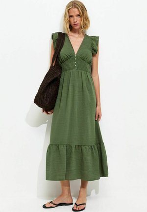 Kobieta w zielonej sukience midi bez rękawów z guzikami i falbaną na dole, niosąca dużą ciemną plecioną torbę typu tote oraz czarne klapki.