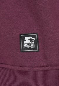 Burgunderfarget hettegenser med strukturiert stoff, med et svart rektangulært merke som har en hvit stjerne og teksten "STARTER BLACK LABEL."