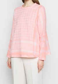 Różowa bluzka z długim rękawem, z wzorem w zęby psa oraz wzorami geometrycznymi, z okrągłym dekoltem i rękawami w kształcie dzwonu. Miękka tkanina o lekko fakturowanym wykończeniu.