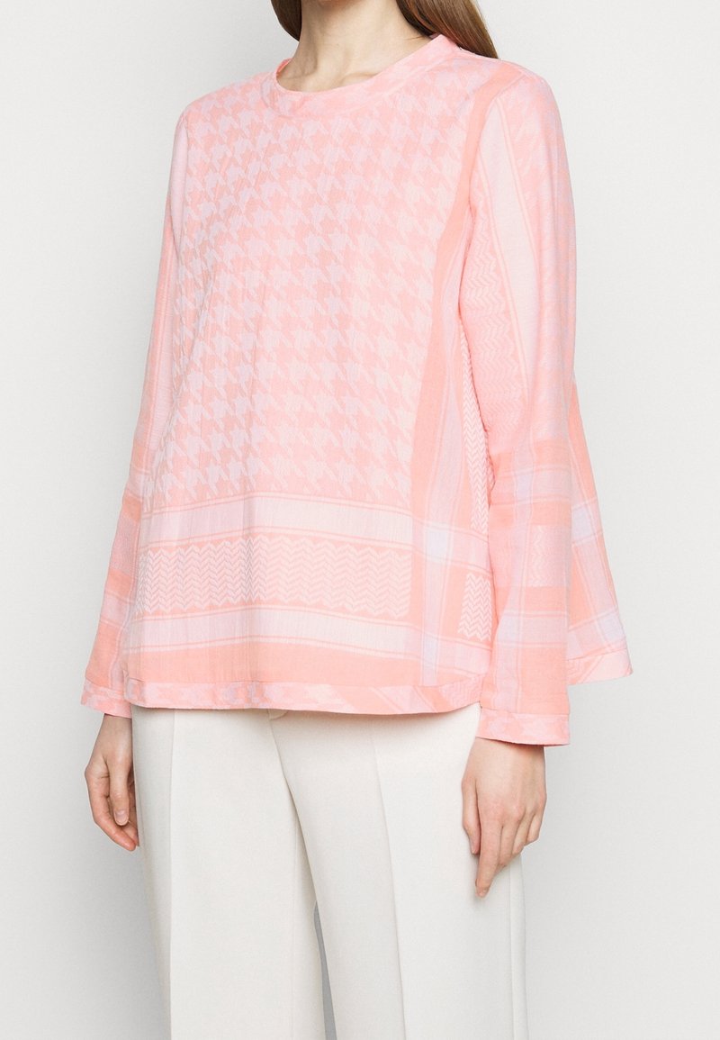 Różowa bluzka z długim rękawem, z wzorem w zęby psa oraz wzorami geometrycznymi, z okrągłym dekoltem i rękawami w kształcie dzwonu. Miękka tkanina o lekko fakturowanym wykończeniu.
