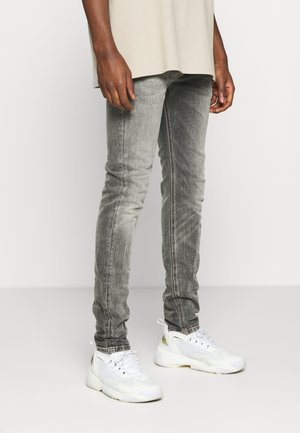 Person, die ausgewaschene graue Skinny-Jeans und weiße Sportschuhe trägt, steht vor einem schlichten weißen Hintergrund.