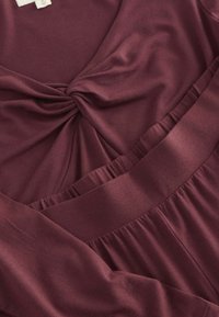 Vestido burdeos con detalle de nudo en la parte delantera, tejido suave, mangas largas y una cintura fruncida con acentos de volantes.
