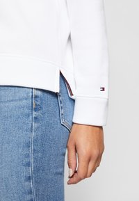 Dešinė asmens ranka, dėvinčio balto ilgomis rankovėmis megztinį su logotipu prie riešo ir mėlynas džinsas.