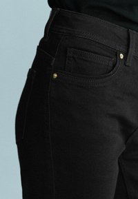 Gros plan d'une personne portant un jean noir ajusté avec des rivets dorés et un haut noir sur un fond clair uni.