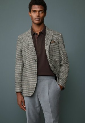Houndstooth-mønsteret blazer i beige og sort, parret med en mørkebrun polo skjorte og lysegrå plissee-bukser.