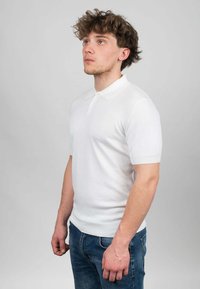 Polo shirt bianco a maniche corte con colletto standard, realizzato in tessuto liscio. Presenta una forma aderente e polsini a coste. Indossato con jeans blu.