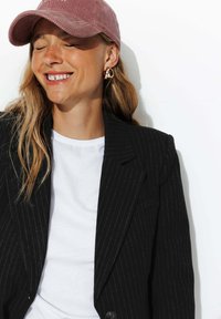 Blazer noir à rayures sur un t-shirt blanc, assorti d'une casquette en velours côtelé rose et de boucles d'oreilles abstraites en or. Le sujet sourit devant un fond blanc.