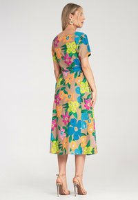 Maxi-Kleid mit floralen Mustern, das einen beigen Hintergrund mit leuchtend blauen, pinken, orangen und grünen Blumen zeigt, kurzen Ärmeln und einem Binderiemen im Rücken.