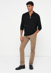 Pullover nero con colletto a zip, abbinato a pantaloni in corduroy beige e mocassini neri. I materiali presentano una texture liscia e una vestibilità su misura.