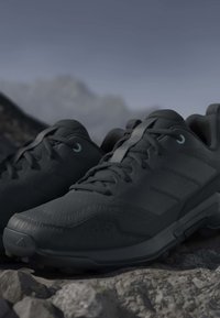 Pair of black athletic shoes with laces on rocky terrain, mountain blurred in background under gray sky.
