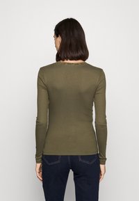 Olivgrünes, langärmeliges, geripptes Oberteil mit rundem Ausschnitt und Spitzenbesatz auf der Rückseite. Weicher, figurbetonter Stoff. Kombiniert mit dunklen Jeans.